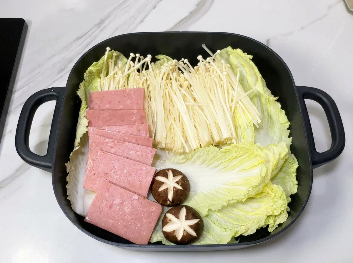 Una pentola quadrata contenente strati ordinati di cavolo cinese, funghi enoki e fette di spam su un letto di cipolle.