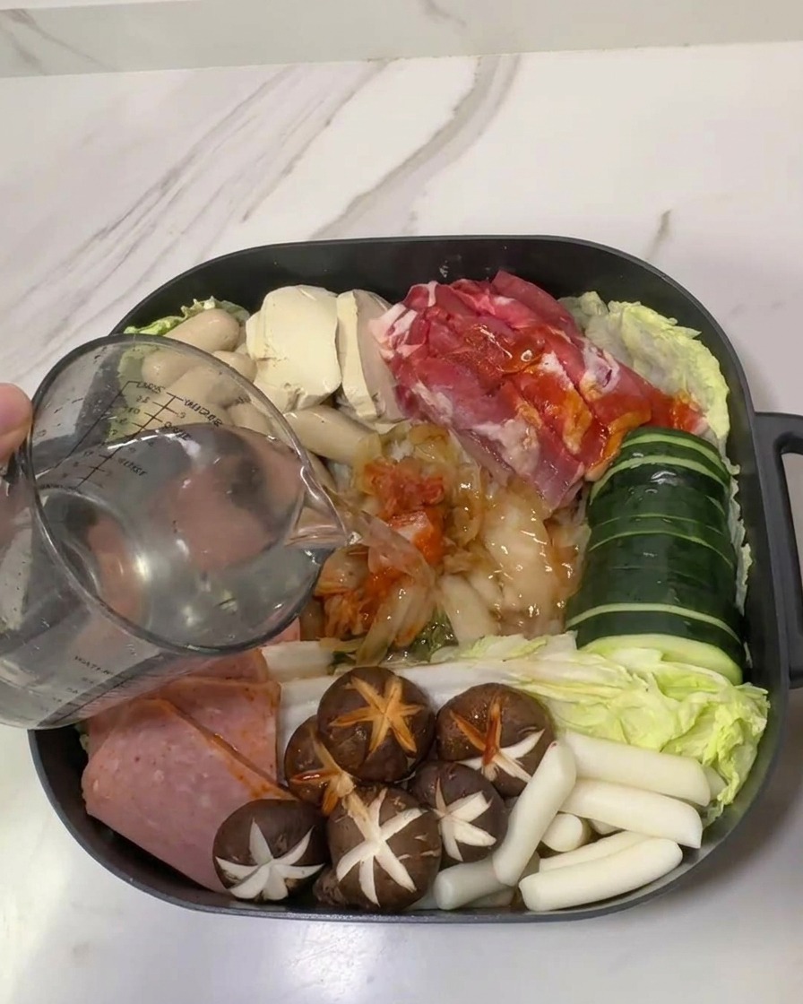Un misurino di vetro che versa acqua in una pentola piena di ingredienti per il Budae Jjigae disposti con cura.