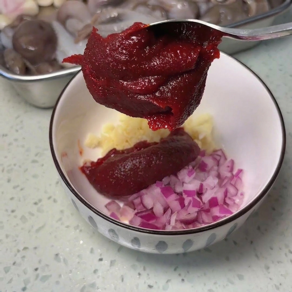 Un cucchiaio di metallo con una densa pasta di peperoncino rosso coreano sopra una ciotola con aglio e cipolla.
