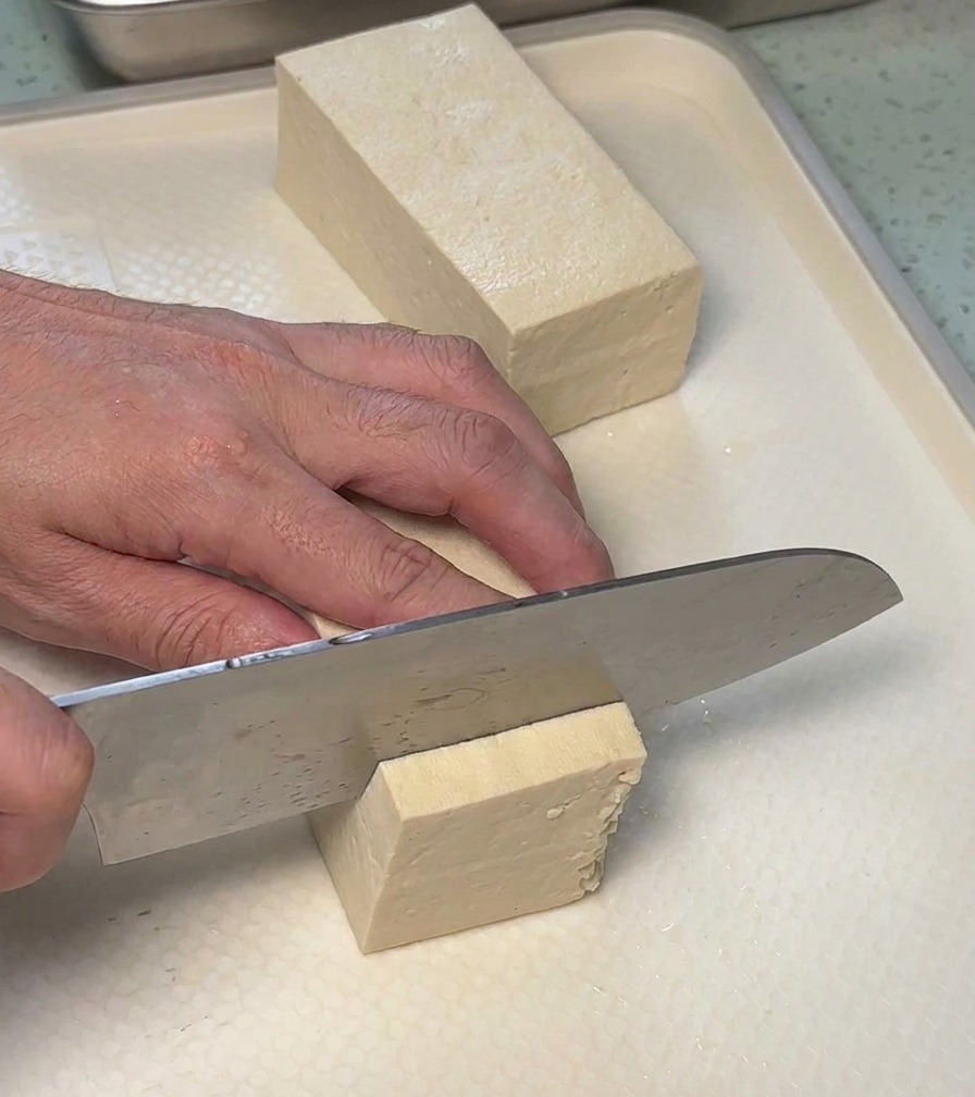 Una persona che taglia un blocco di tofu bianco a fette rettangolari su un tagliere bianco.