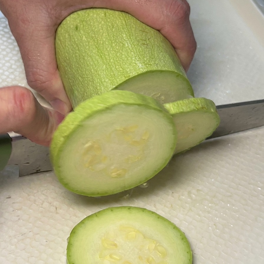 Mani che tagliano una zucchina verde chiaro a rondelle su un tagliere bianco.