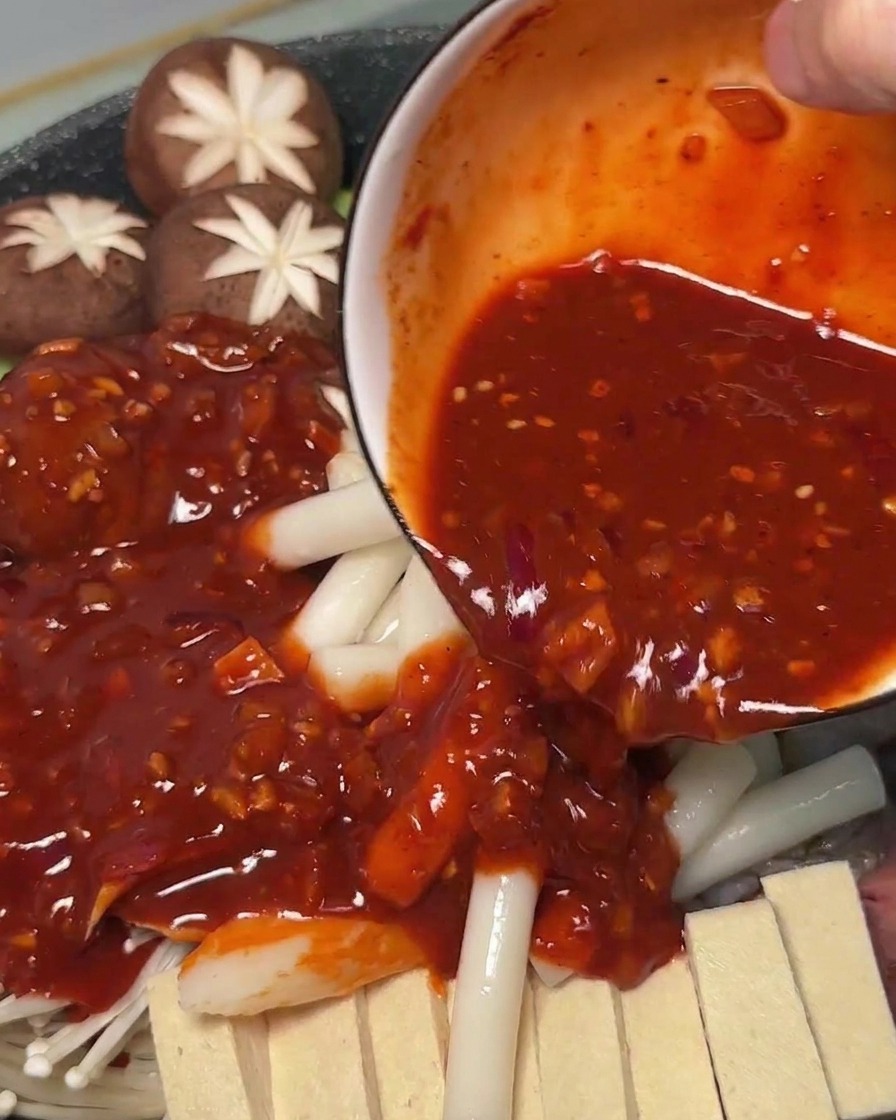 Salsa rossa piccante versata da una ciotola sugli ingredienti crudi e gli gnocchi di riso nella pentola.