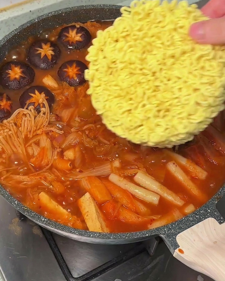 Una mano che mette un panetto di ramen istantaneo in una pentola di zuppa rossa bollente.