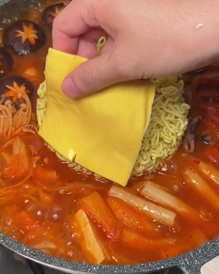 Una fetta di formaggio giallo posizionata sopra i noodles in una pentola di Budae Jjigae bollente.