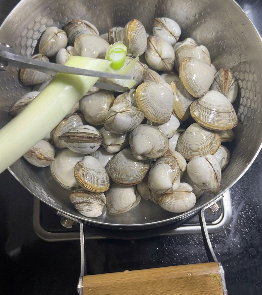 Una mano che usa le forbici per tagliare un cipollotto sopra una padella con vongole bianche e fette di zenzero.
