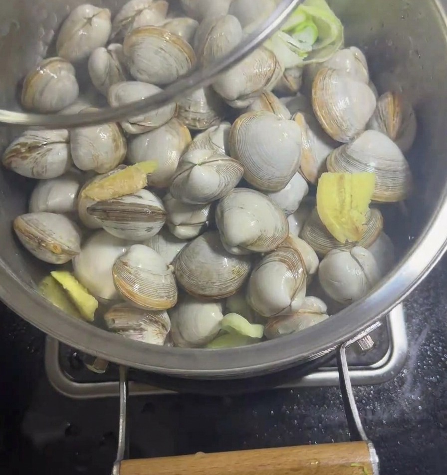 Una padella in acciaio inox con vongole e aromi che viene coperta con un coperchio di vetro trasparente.
