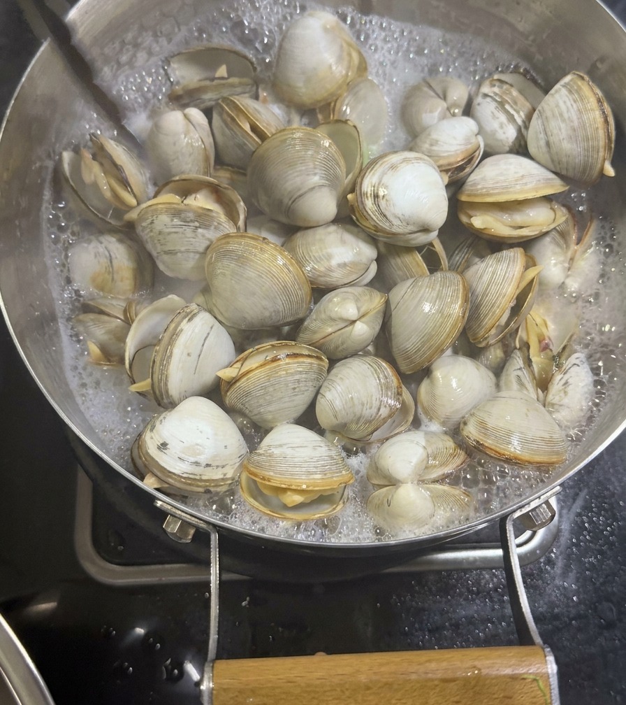 Pinze di metallo che sollevano vongole bianche aperte da una padella con un brodo di mare lattiginoso e bollente.
