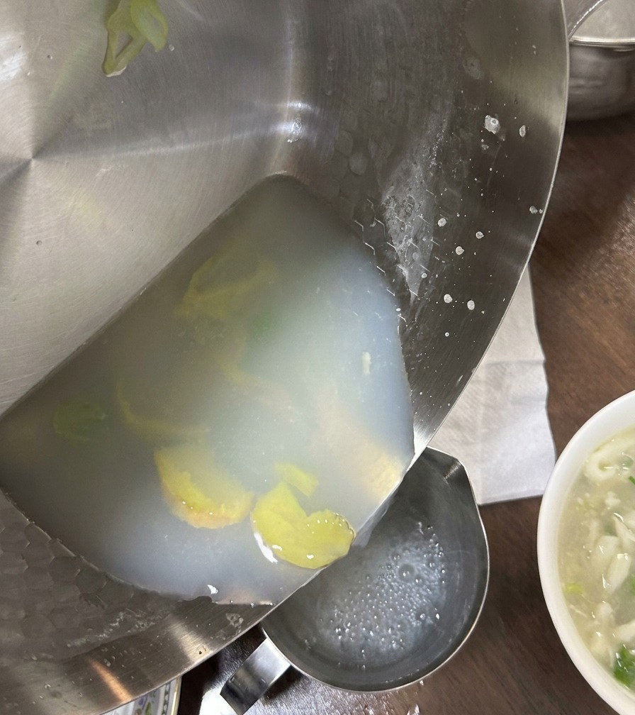 Brodo di vongole bianco lattiginoso versato da una padella in un contenitore, lasciando indietro lo zenzero e i residui.