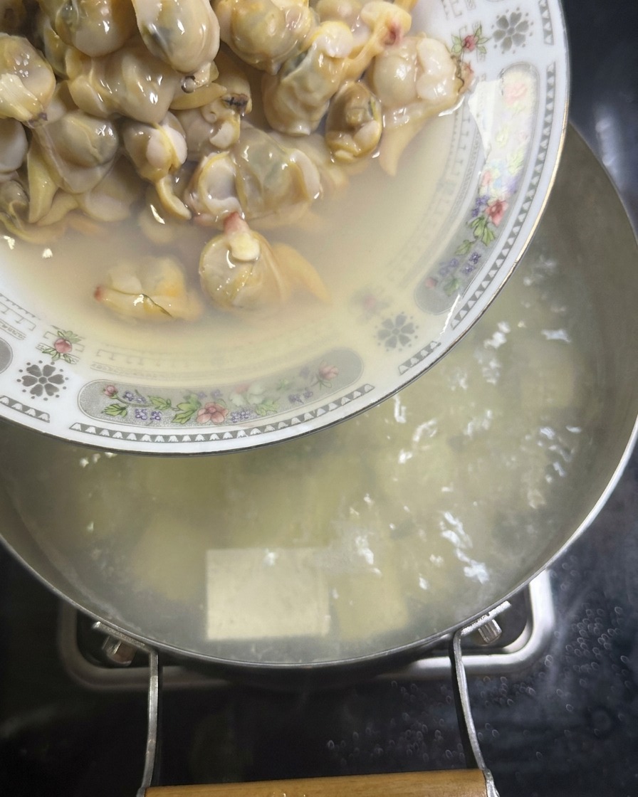 Un piatto di polpa di vongole cotta che viene versato in una pentola di brodo bianco con cubetti di tofu.