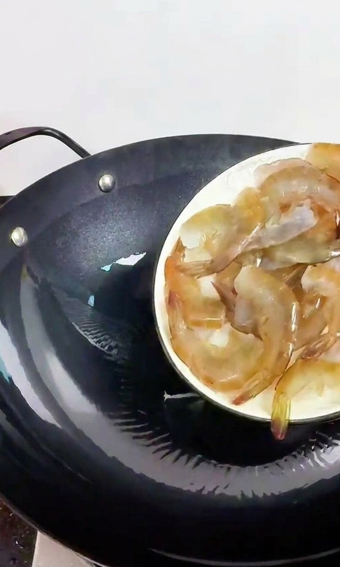 Una mano che tiene un piatto bianco, facendo scivolare gamberi freschi e traslucidi in un wok di ferro nero preriscaldato.