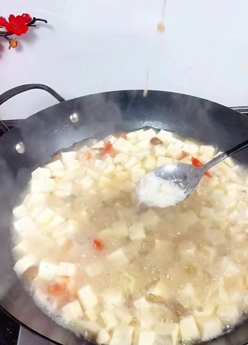 Un cucchiaio di metallo che sparge sale bianco in un grande wok pieno di cubetti di tofu e frutti di mare in un brodo leggero.