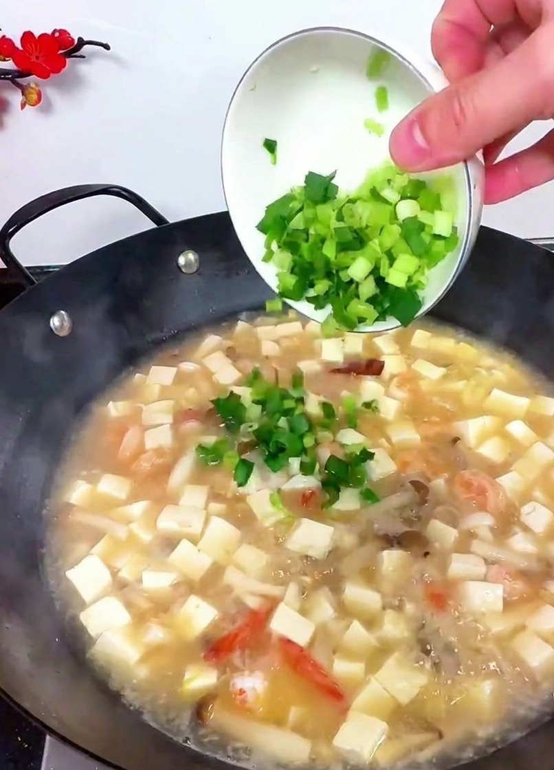Una mano che inclina una piccola ciotola bianca per versare sedano verde brillante e germogli daglio tritati in un wok di zuppa di tofu bollente.