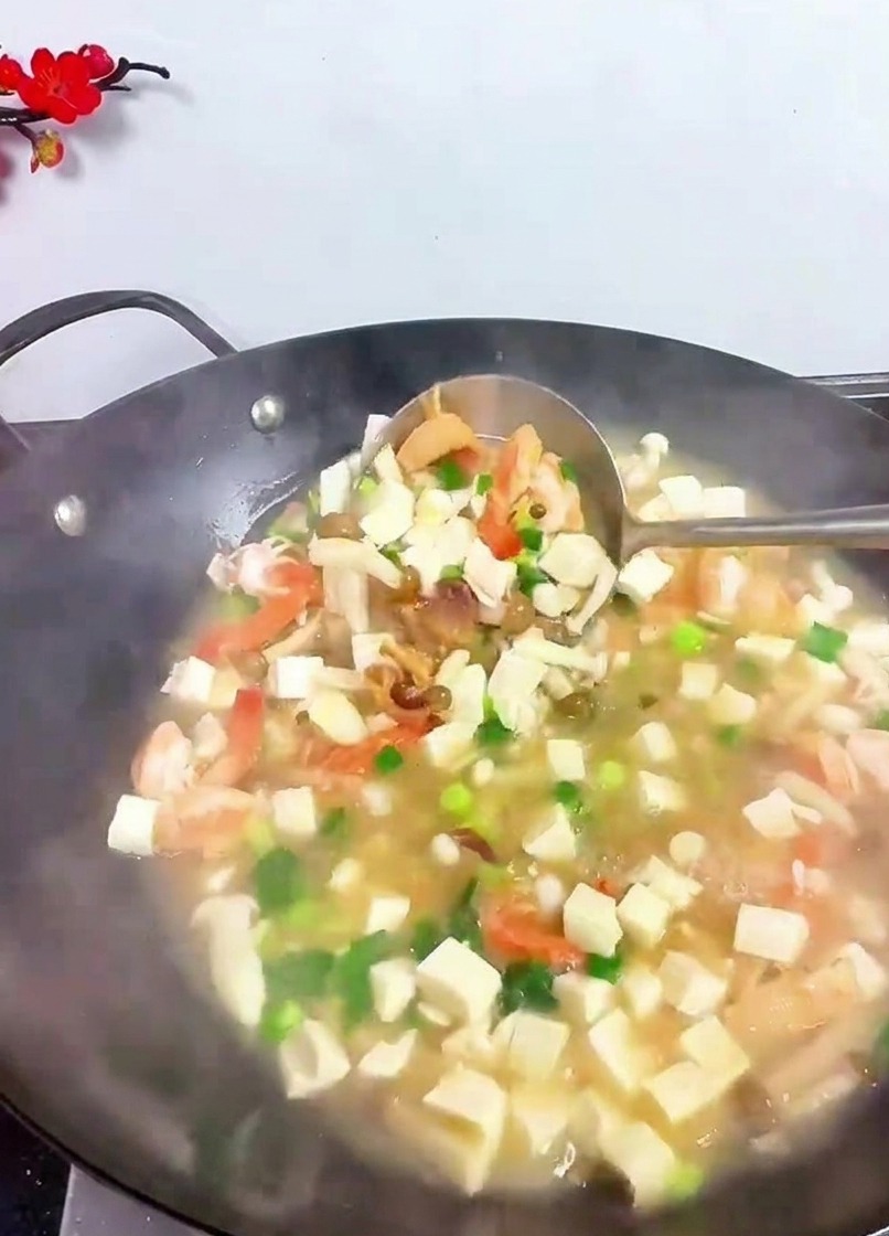Un mestolo di metallo che solleva e mescola vari ingredienti tra cui cubetti di tofu, gamberi ed erbe verdi in un wok fumante.