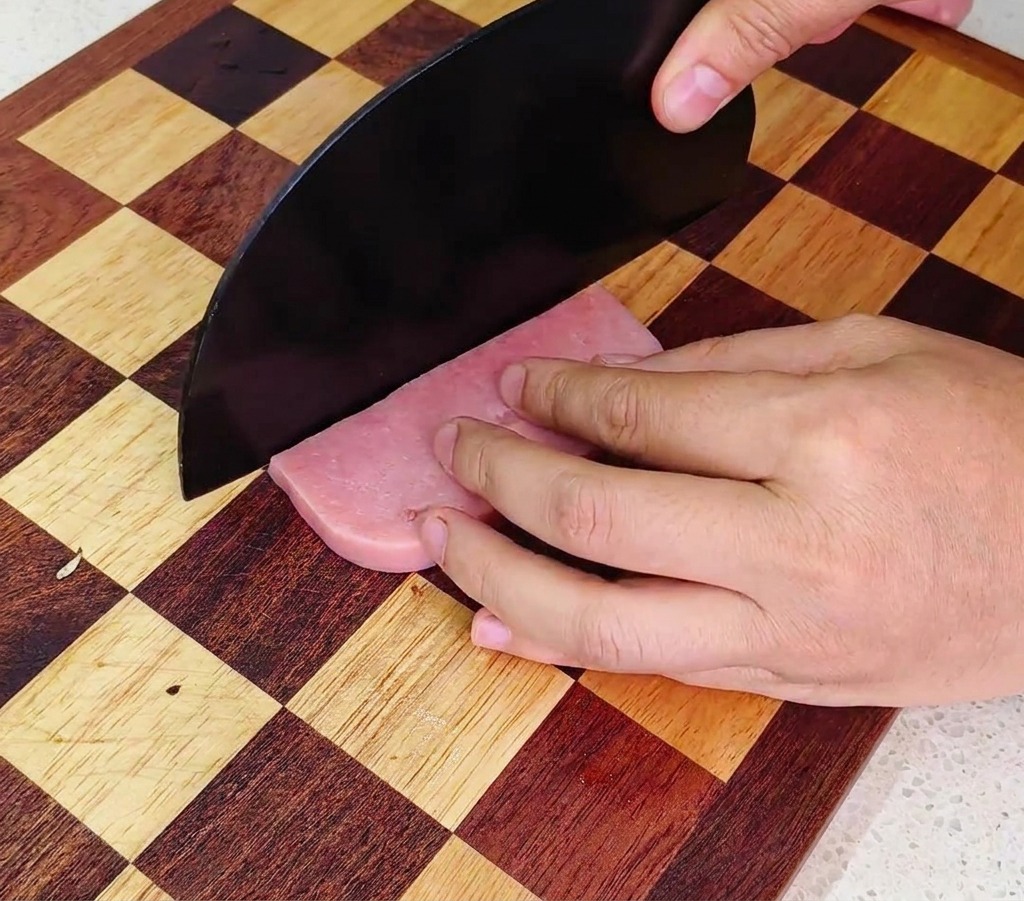 Una persona utilizza un coltello da chef per tagliare il prosciutto a cubetti uniformi su un tagliere di legno.