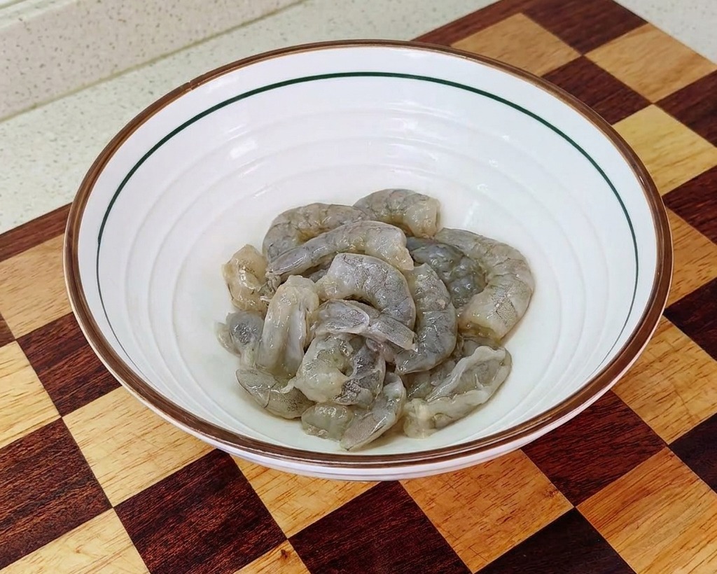 Una ciotola bianca contenente gamberi puliti conditi con sale, amido e olio per la marinatura.
