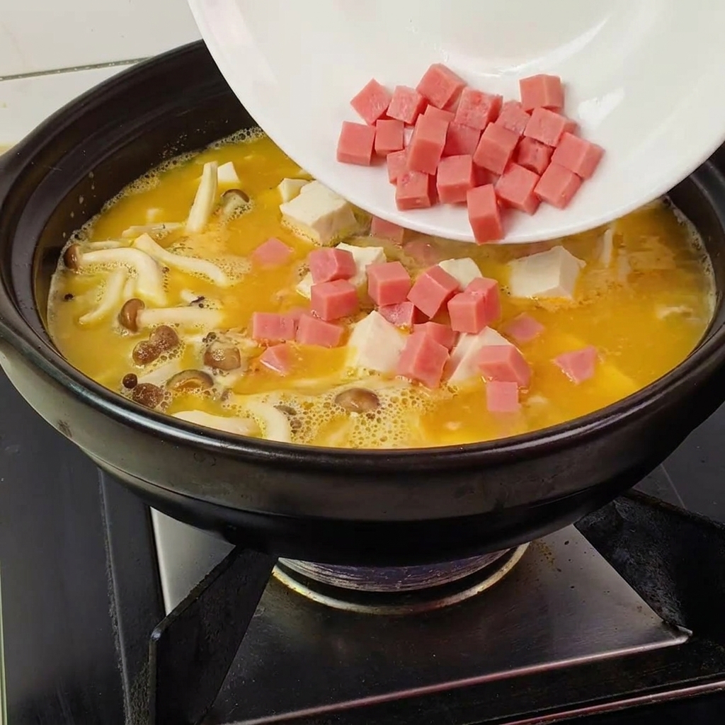 Prosciutto a cubetti versato in una pentola con brodo giallo, funghi e tofu.