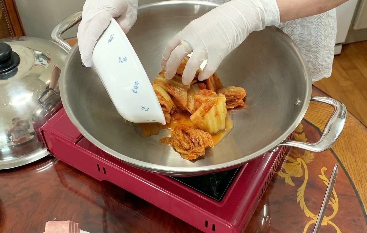 Una persona aggiunge il kimchi affettato da una ciotola al centro di una grande pentola dacciaio su un fornello portatile.