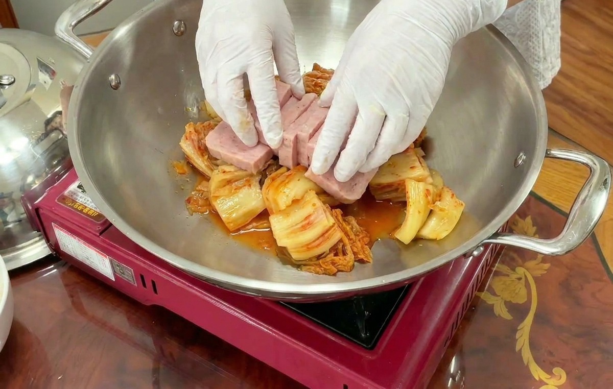 Mani che sistemano fette rettangolari di carne Spam sopra un letto di kimchi in una pentola dacciaio.