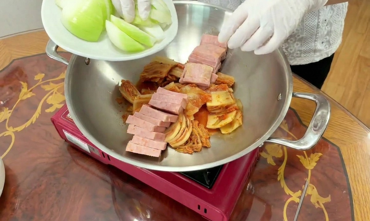 Vengono aggiunti spicchi di cipolla bianca fresca in una pentola che contiene già kimchi e Spam affettato.