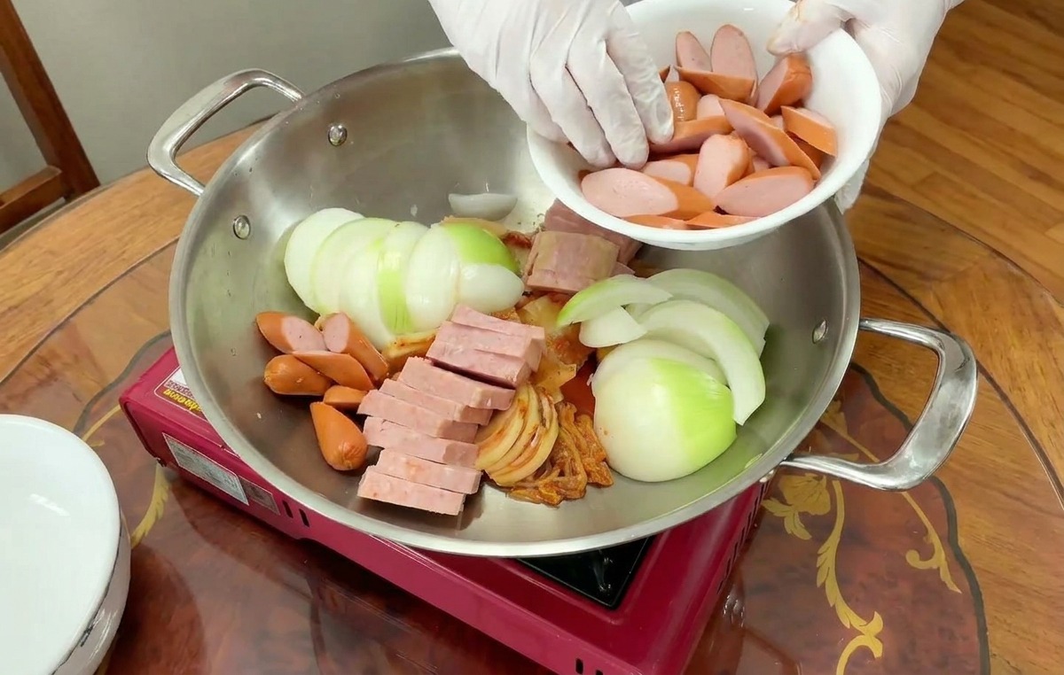Salsicce affettate versate da una ciotola bianca in una pentola con cipolle, carne e kimchi.