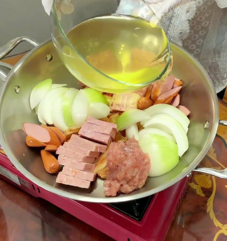 Liquido giallo limpido versato da una ciotola di vetro in una pentola dacciaio piena di ingredienti crudi.