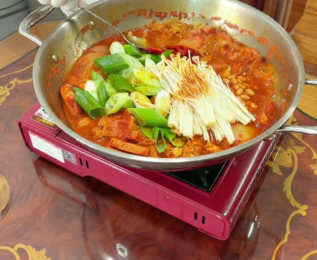 Mani che usano un cucchiaio per mescolare una pentola bollente di Budae Jjigae, amalgamando carne e verdure col brodo rosso.