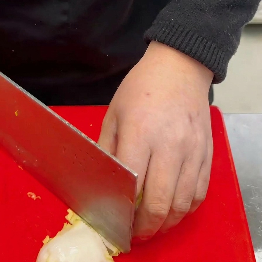 Una mano che usa una grande mannaia per tagliare un cespo di cavolo baby bianco in pezzi grossolani su un tagliere rosso.