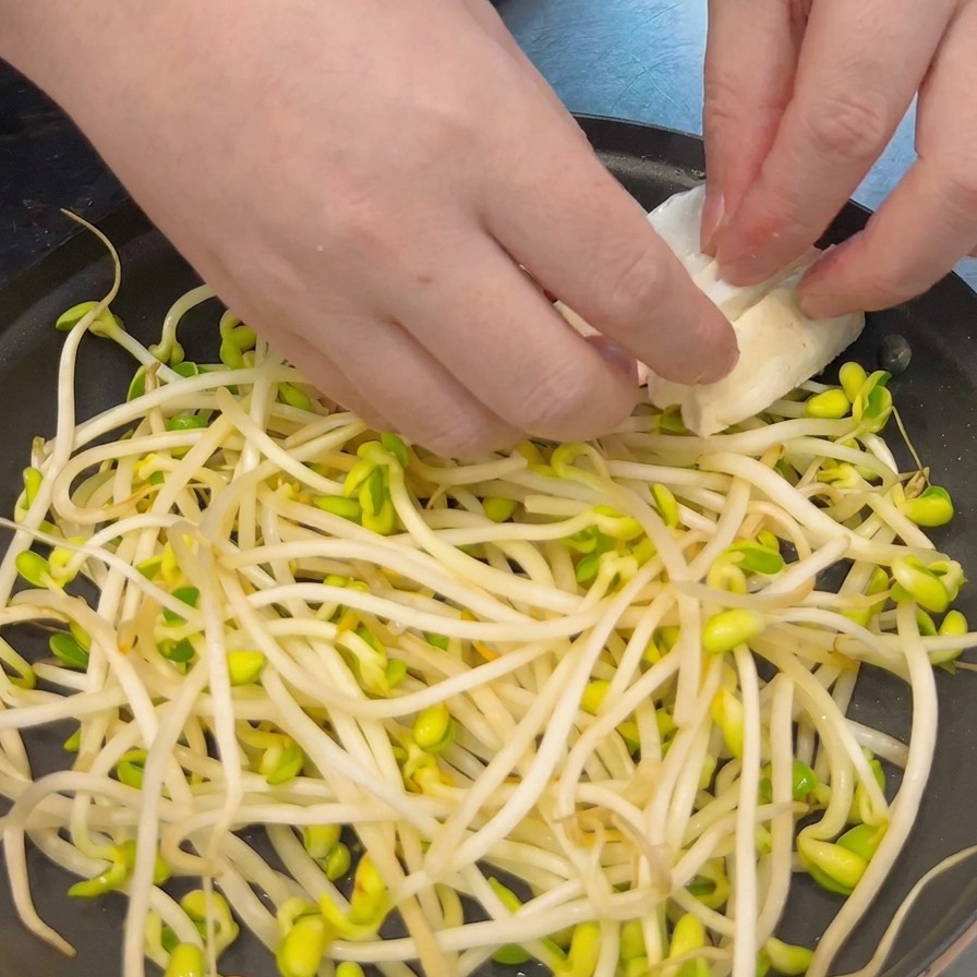 Mani che posizionano con cura fette spesse di tofu bianco su un letto di germogli di soia in una pentola nera bassa.