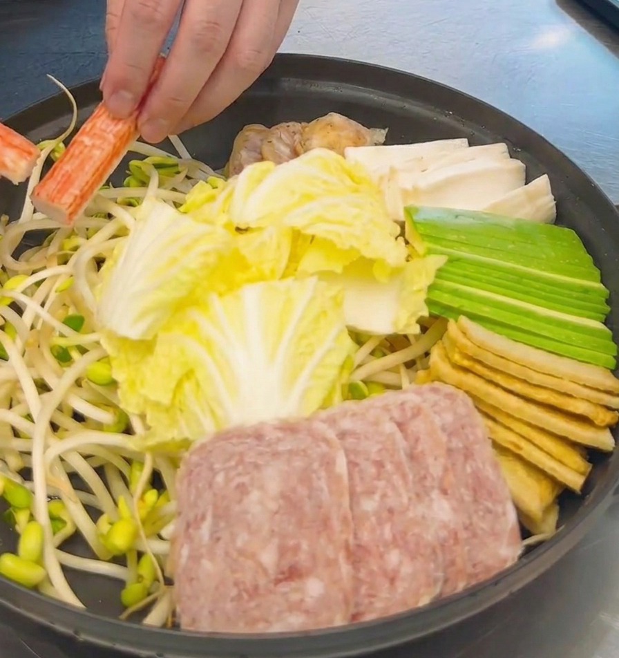 Una mano che dispone bastoncini di granchio arancioni e bianchi in una pentola già riempita con cavolo affettato, spam e tofu.