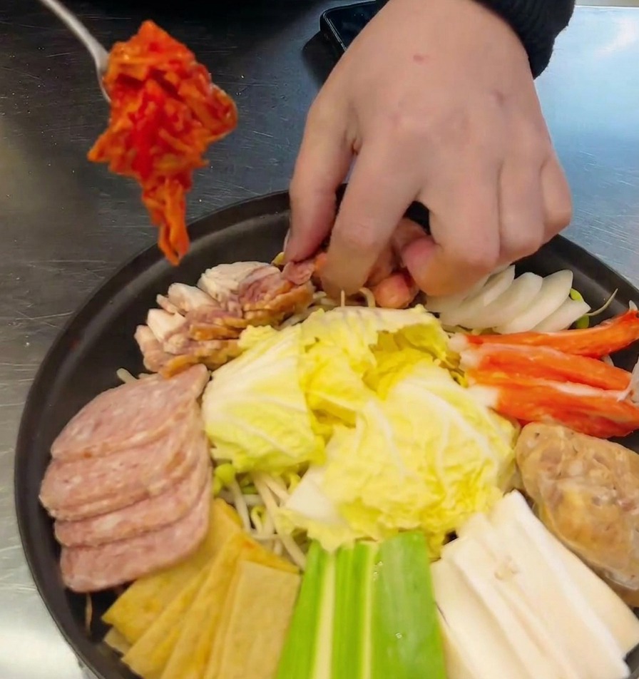 Una mano che usa un cucchiaio di metallo per mettere una porzione di kimchi rosso in una pentola bassa con salsicce, spam e verdure.