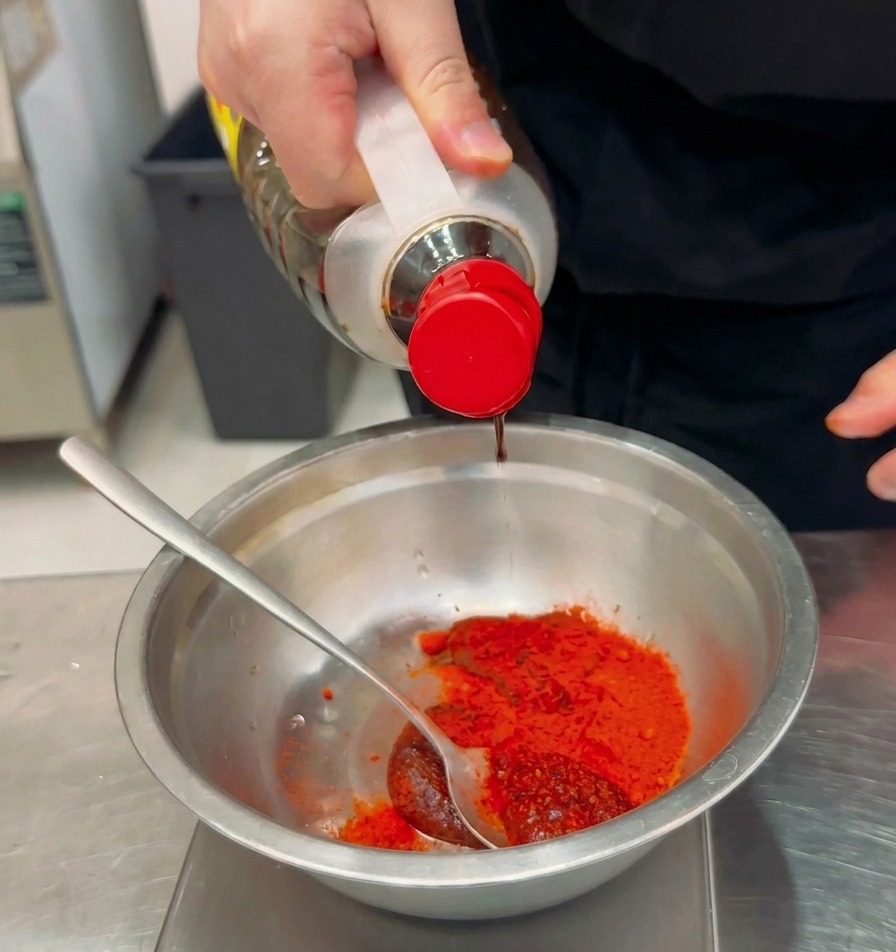 Condimento liquido versato da una bottiglia trasparente in una ciotola di metallo contenente peperoncino in polvere e pasta rossa.