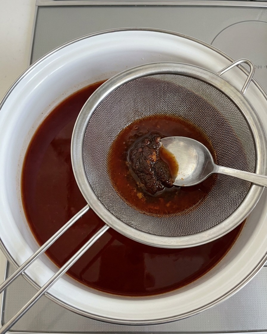 Un cucchiaio che preme la pasta di soia attraverso un colino metallico nel brodo bollente.