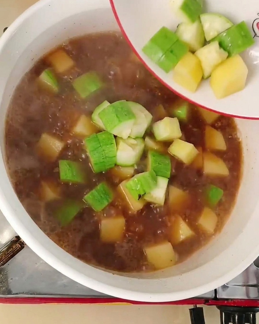 Zucchine a cubetti e cipolle versate da un piatto bianco in un brodo bollente rossastro.