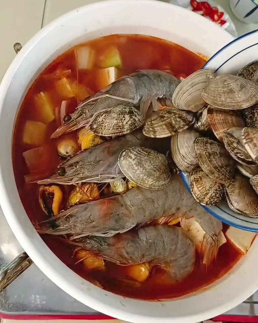 Vongole fresche versate da una ciotola blu in una pentola colma di gamberi e tofu.