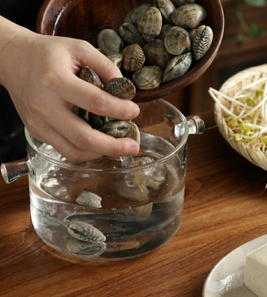 Mani che versano vongole fresche in una ciotola di vetro con acqua su un tavolo di legno.
