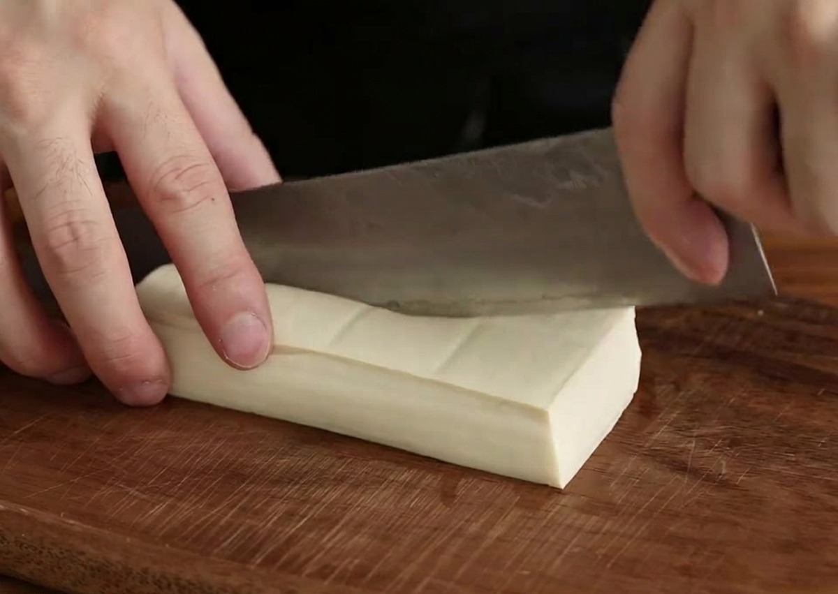 Mano con un coltello da chef che taglia un blocco di tofu bianco in cubetti su un tagliere di legno scuro.