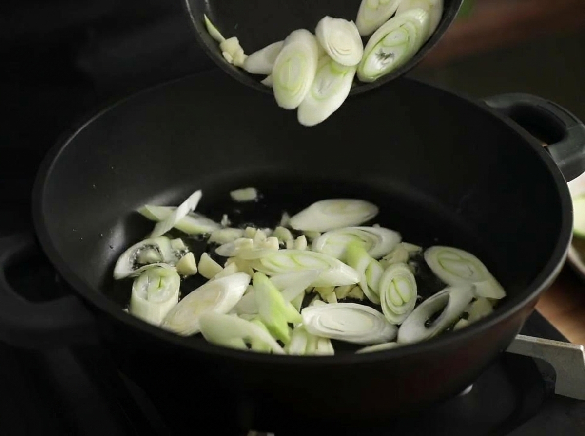 Aglio tritato e fette di porro che soffriggono in una pentola nera con olio lucente.