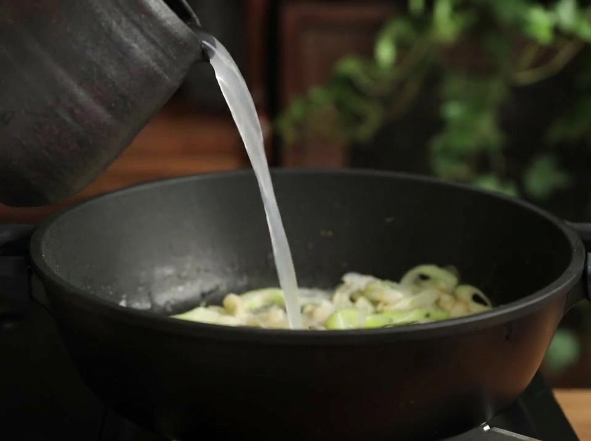 Acqua di riso torbida versata da una brocca in una pentola con porri e aglio soffritti.