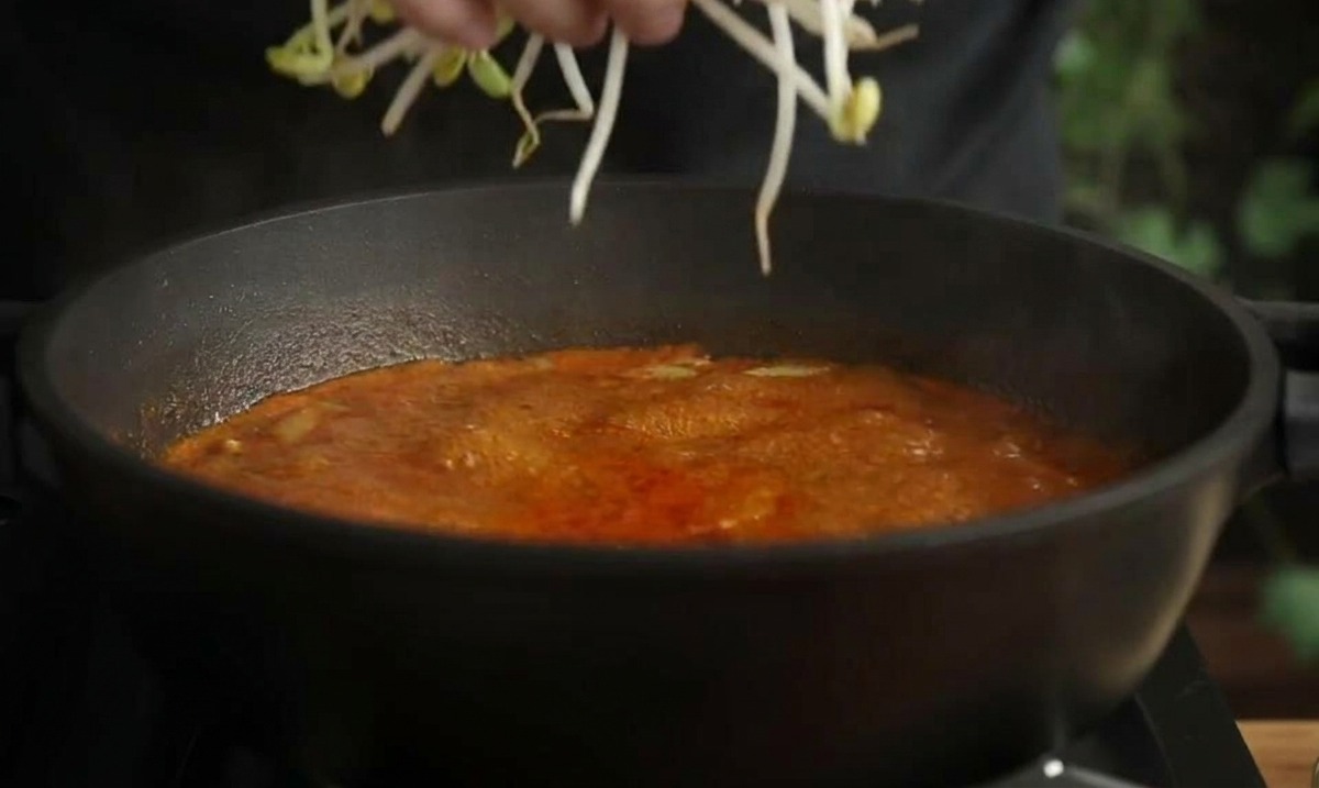 Mano che aggiunge una manciata di germogli di soia lunghi in una pentola di zuppa di pesce rossa bollente.