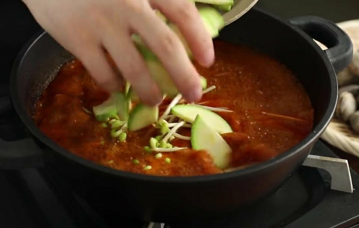 Mani che mettono fette di zucchina fresca nel brodo rosso bollente che già contiene i germogli.