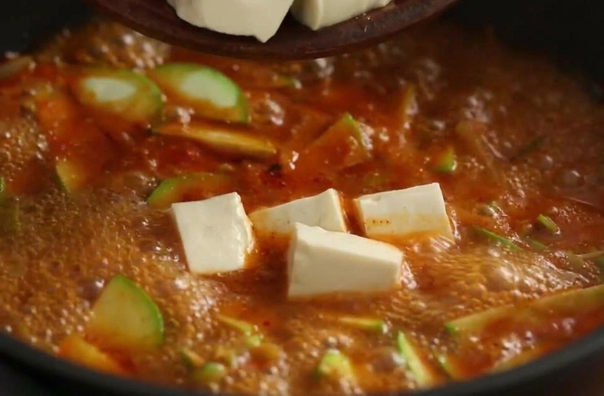 Cubetti di tofu bianco versati da una ciotola in uno stufato coreano rosso e bollente.