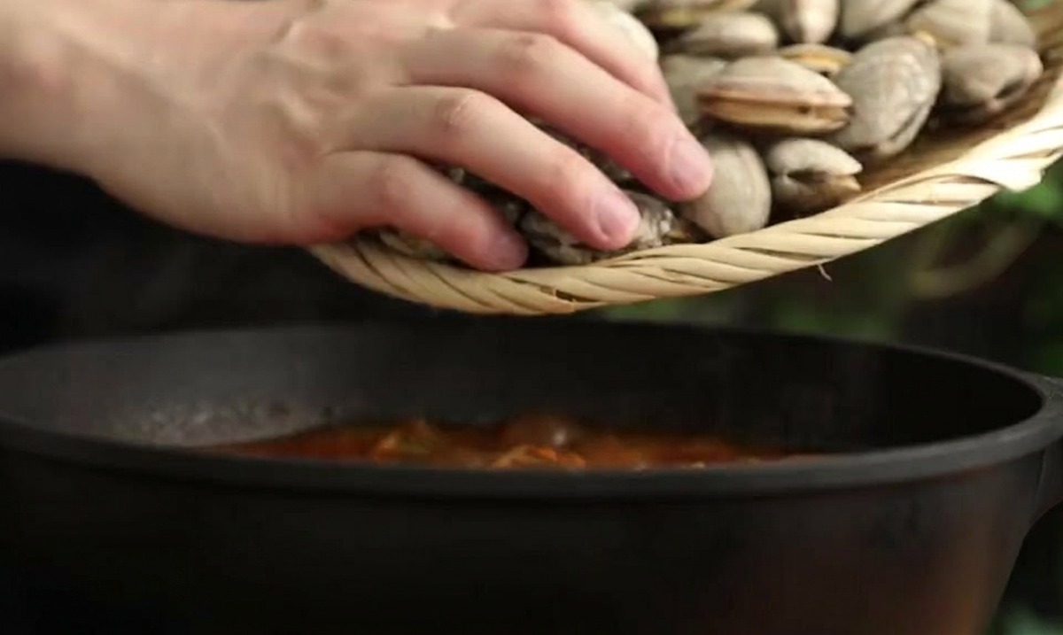 Persona che usa un colino di bambù per immergere vongole fresche in una pentola scura di stufato rosso.