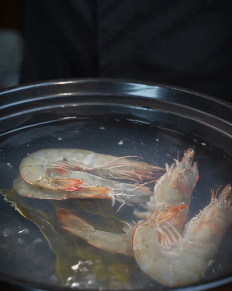 Vongole fresche versate da una ciotola in una pentola che già contiene gamberi e brodo.