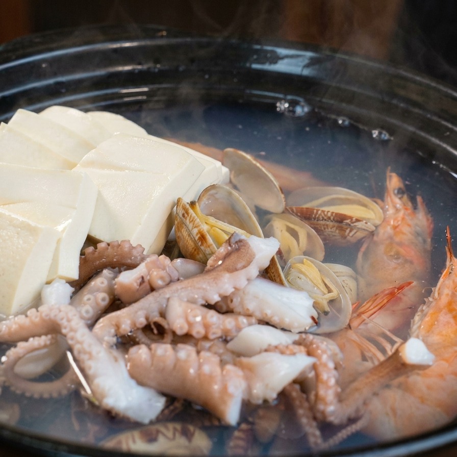 Una pentola di terracotta con vongole e gamberi a cui vengono aggiunte fette di ravanello bianco e carote arancioni.