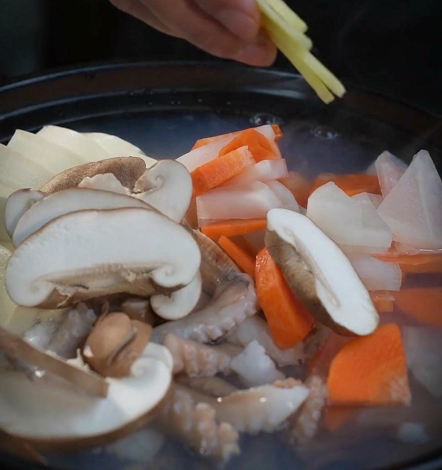 Una mano che posiziona sottili fette di zenzero fresco in una pentola bollente con funghi e frutti di mare.