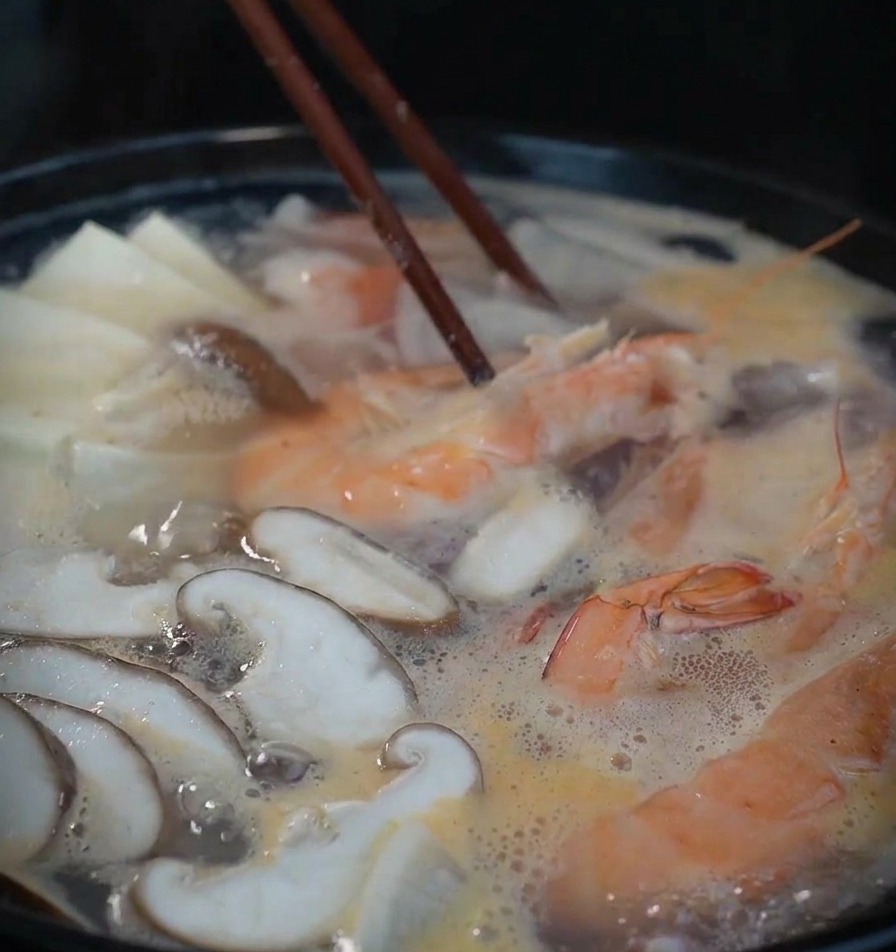 Una pentola nera fumante con gamberi, funghi e tofu che sobbolle, mescolata con bacchette di legno scuro.