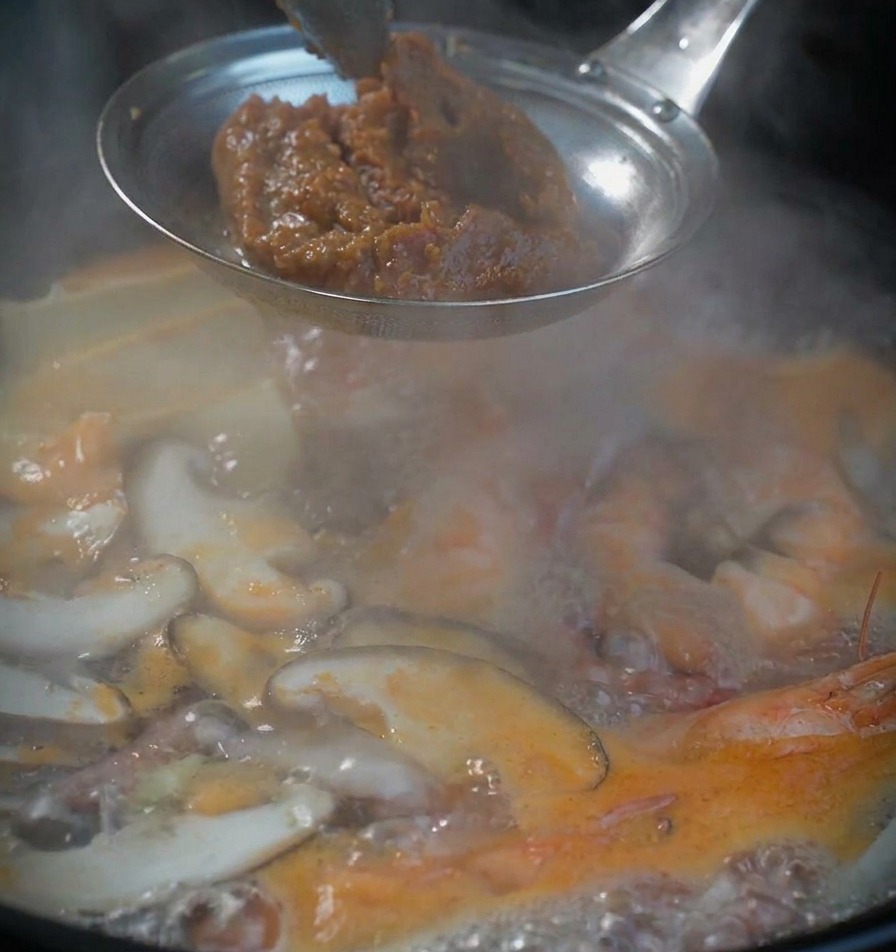Una cucchiaiata di densa pasta di soia marrone in un colino di metallo sopra la pentola della zuppa.