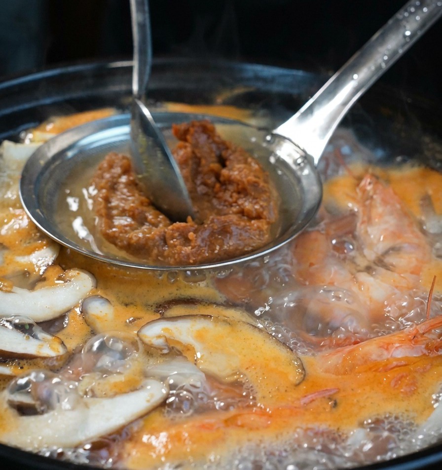 Una mano che usa un cucchiaio per premere la pasta di soia attraverso un colino nel brodo bollente.