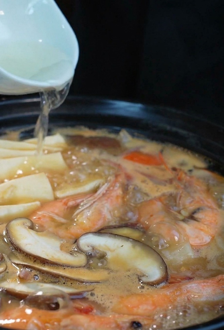 Un filo di sake limpido versato da una ciotolina in una pentola nera fumante con pesce e funghi.