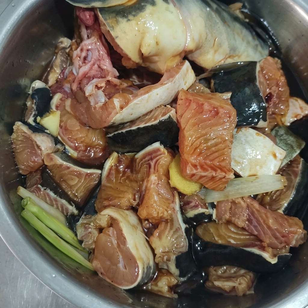 Pezzi di pesce crudo mescolati con zenzero, cipollotti e marinata scura in una ciotola di metallo.